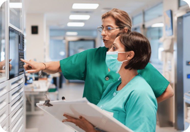 Two healthcare workers in scrubs review information on a screen in a medical facility. One points at the screen, while the other holds a clipboard and wears a face mask.