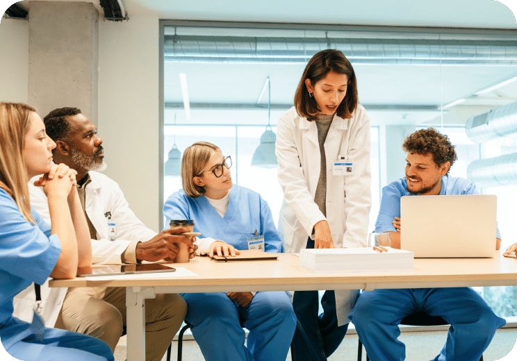 Five healthcare professionals in scrubs and lab coats sit at a table in discussion. One woman stands, pointing at documents, while others listen and take notes. A laptop and coffee cup are on the table.