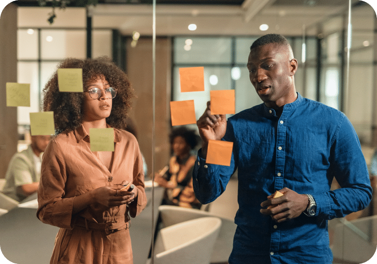 Two people stand by a glass wall covered with sticky notes, brainstorming in a modern office. The man in blue gestures while the woman listens. Other colleagues are visible in the blurred background.
