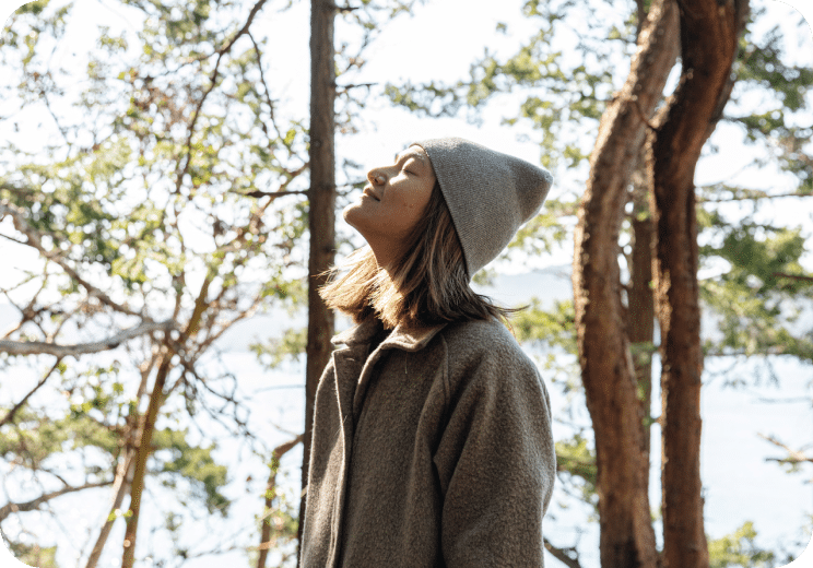 A person in a gray coat and beanie stands outdoors among trees, eyes closed, facing sunlight, appearing calm and peaceful.