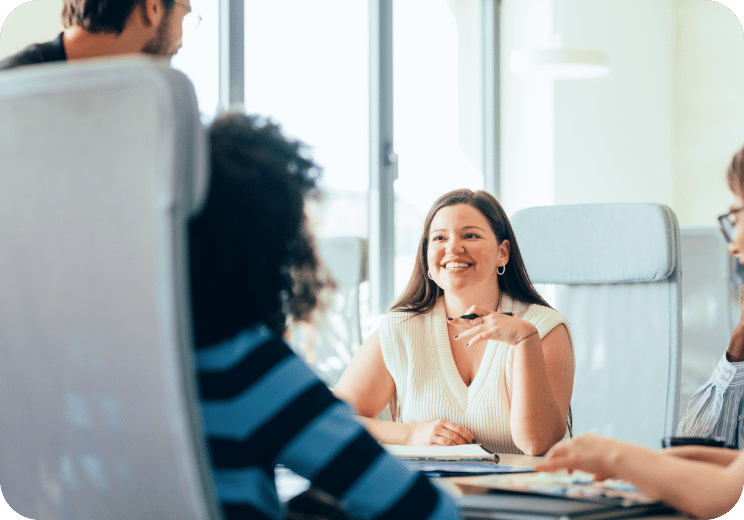 A woman smiles while sitting at a table with three colleagues in a bright, modern office setting. The group appears to be engaged in a friendly discussion or meeting.