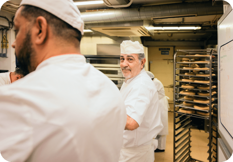 A baker in a white uniform and hat smiles at the camera in a busy bakery kitchen, with trays of bread in the background and another worker in the foreground.