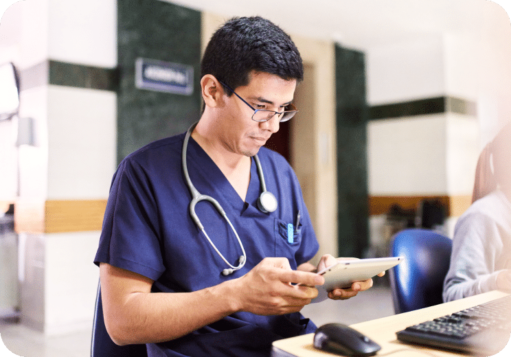 A healthcare professional wearing navy scrubs and a stethoscope looks at a tablet while seated at a desk in a medical facility.