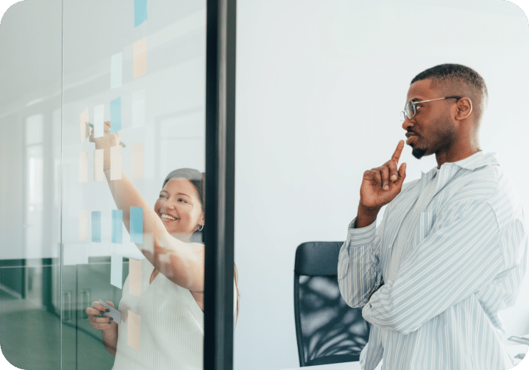 Two people brainstorm in an office, one writing colorful sticky notes on a glass wall while the other stands nearby, thinking and observing the notes.