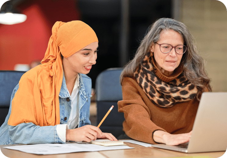 Two women sit at a table working together. One wears a headscarf and holds a pencil, while the other wears glasses and a leopard-print scarf, using a laptop. Papers and pens are on the table.