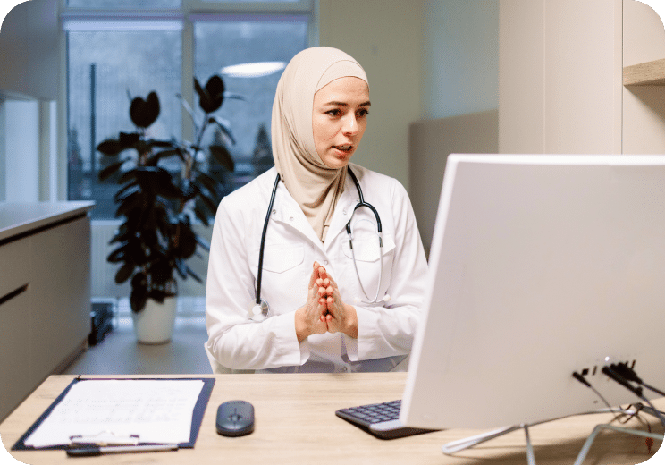 A female doctor in a white coat and hijab sits at a desk with a computer, clipboard, and stethoscope around her neck, appearing to speak during a virtual consultation in a modern office.