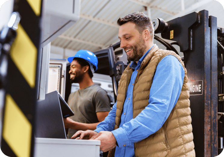 Two men working in a warehouse; one types on a computer while smiling, and the other, wearing a blue hard hat, operates machinery in the background.