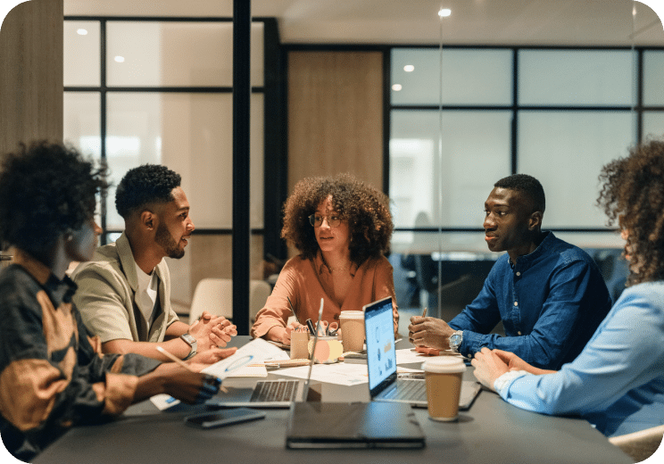 Five people sit around a conference table in a modern office, engaged in discussion. Laptops, papers, and coffee cups are on the table, and a windowed wall is in the background.