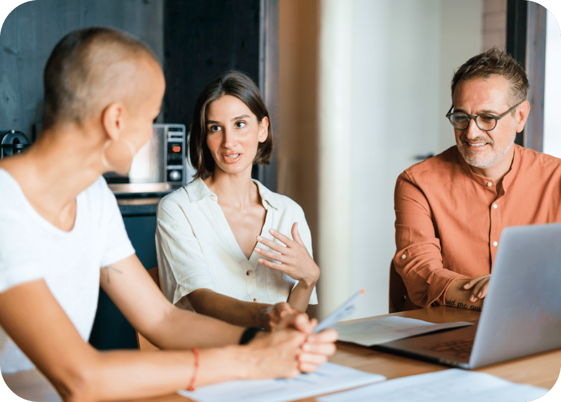Three people sit at a table with papers and a laptop, engaged in discussion. One person speaks while the others listen attentively.