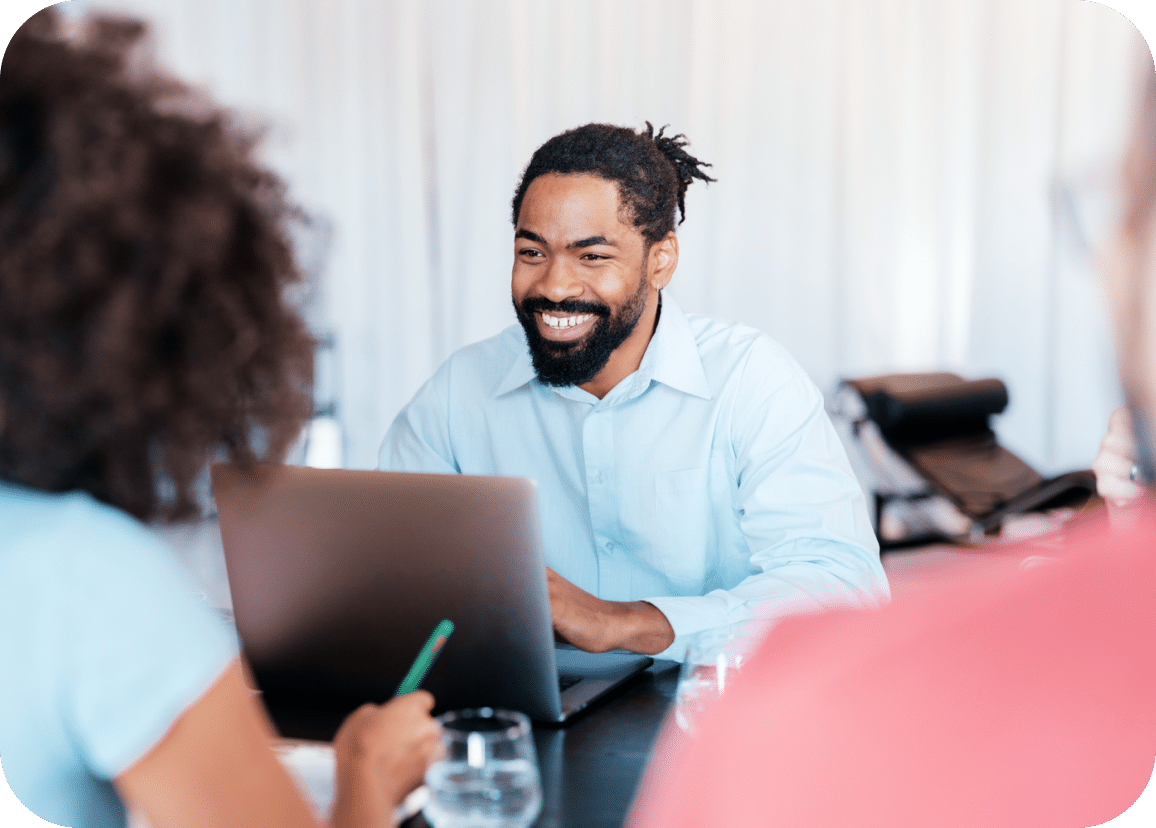 A man with a beard and mustache smiles while working on a laptop at a table, with two blurred people in the foreground.