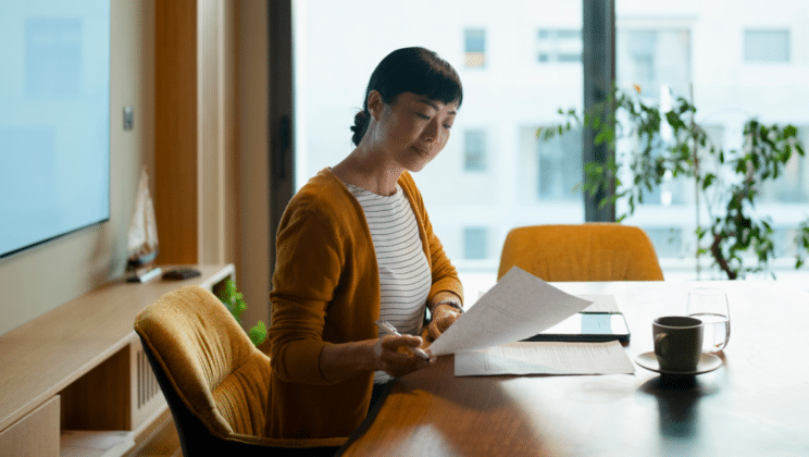 Woman sitting at a table in a modern office, reading documents with a pen in hand. A tablet, cup of coffee, and glass of water are on the table. Large window with city view and a potted plant in the background.