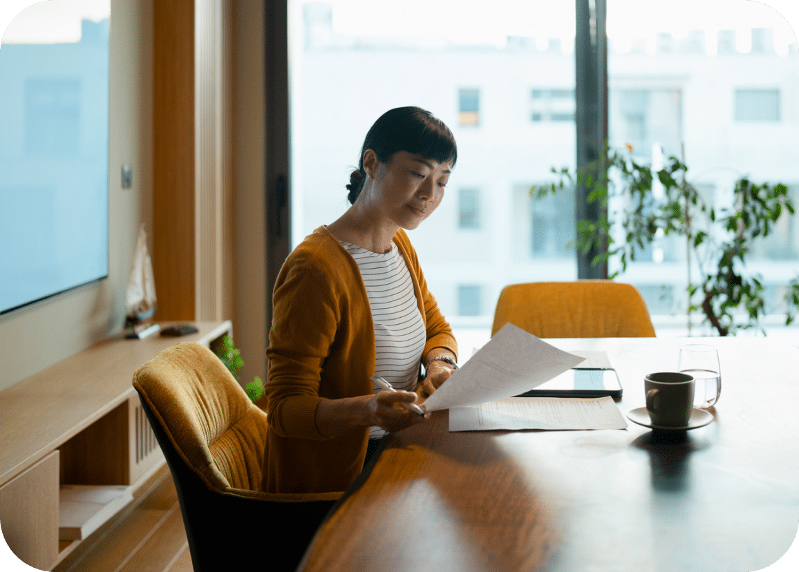 Woman sitting at a table in a modern office, reading documents with a pen in hand. A tablet, cup of coffee, and glass of water are on the table. Large window with city view and a potted plant in the background.