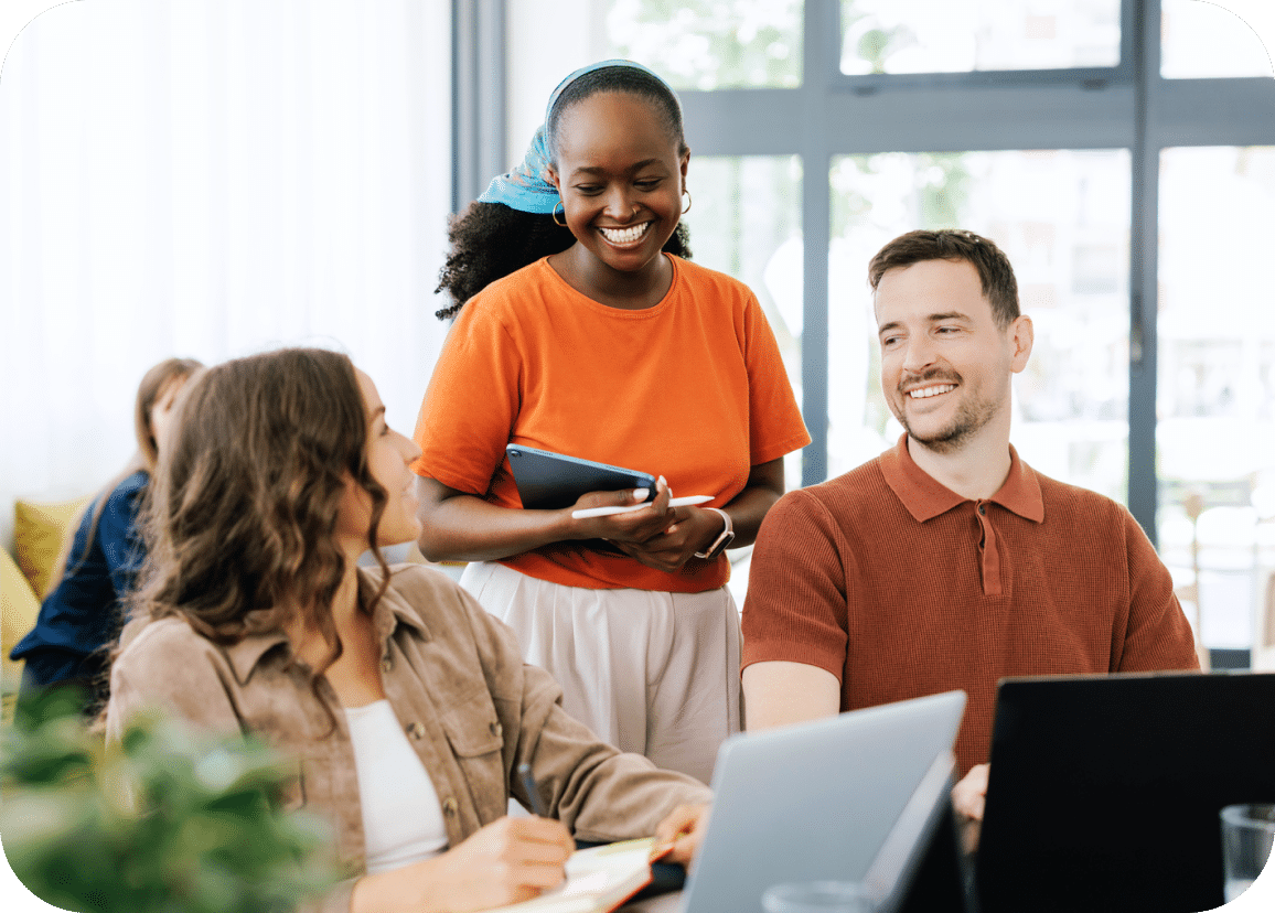 Three people in a bright office; two sit at laptops while one stands, holding a tablet and smiling, as they interact.