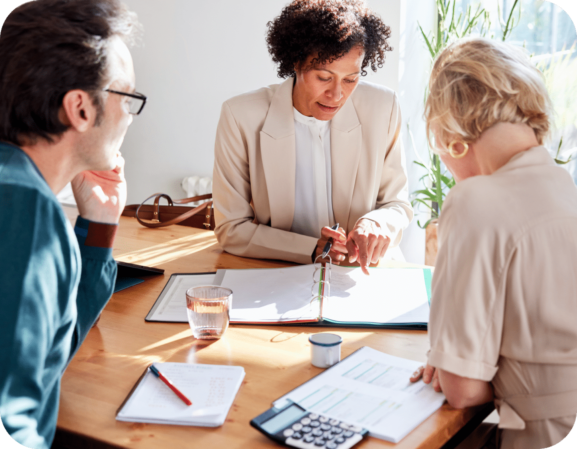 Three people sit at a wooden table with documents, a calculator, and a glass of water, discussing paperwork. One woman points at a binder while the others listen and take notes. Sunlight streams in from the side.