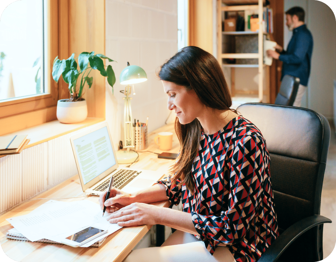 A woman works at a desk with papers, a laptop, and a phone. She writes with a pen while sitting near a window. A man stands in the background by an open closet or shelf. The workspace looks bright and organized.