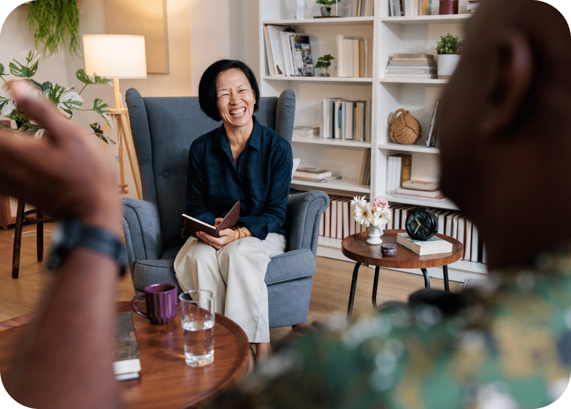 A smiling woman sits in an armchair holding a notebook, listening to someone speaking. They are in a cozy room with bookshelves, plants, and coffee tables.