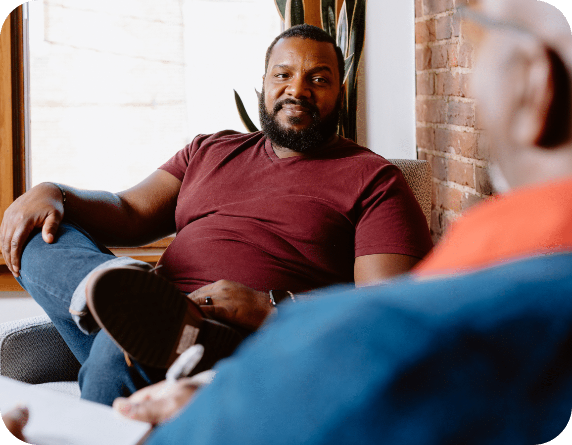 A man in a maroon shirt sits on a couch with his leg crossed, smiling and talking to another person whose back is to the camera. They are in a cozy room with a brick wall and large window.