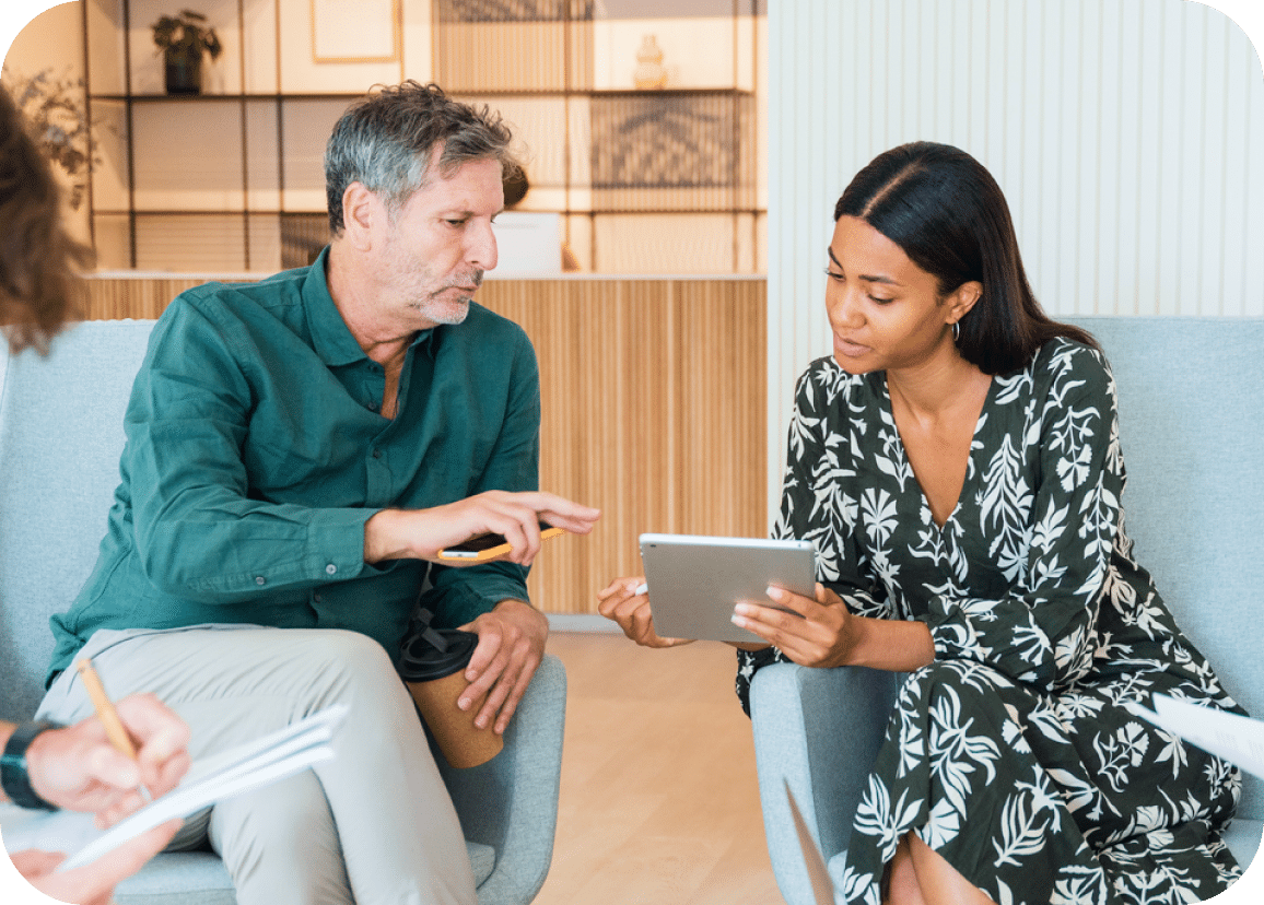 Two people sit in chairs, discussing something displayed on a tablet. The man gestures towards the screen while the woman holds the device. Both appear focused, collaborating in a modern office setting.