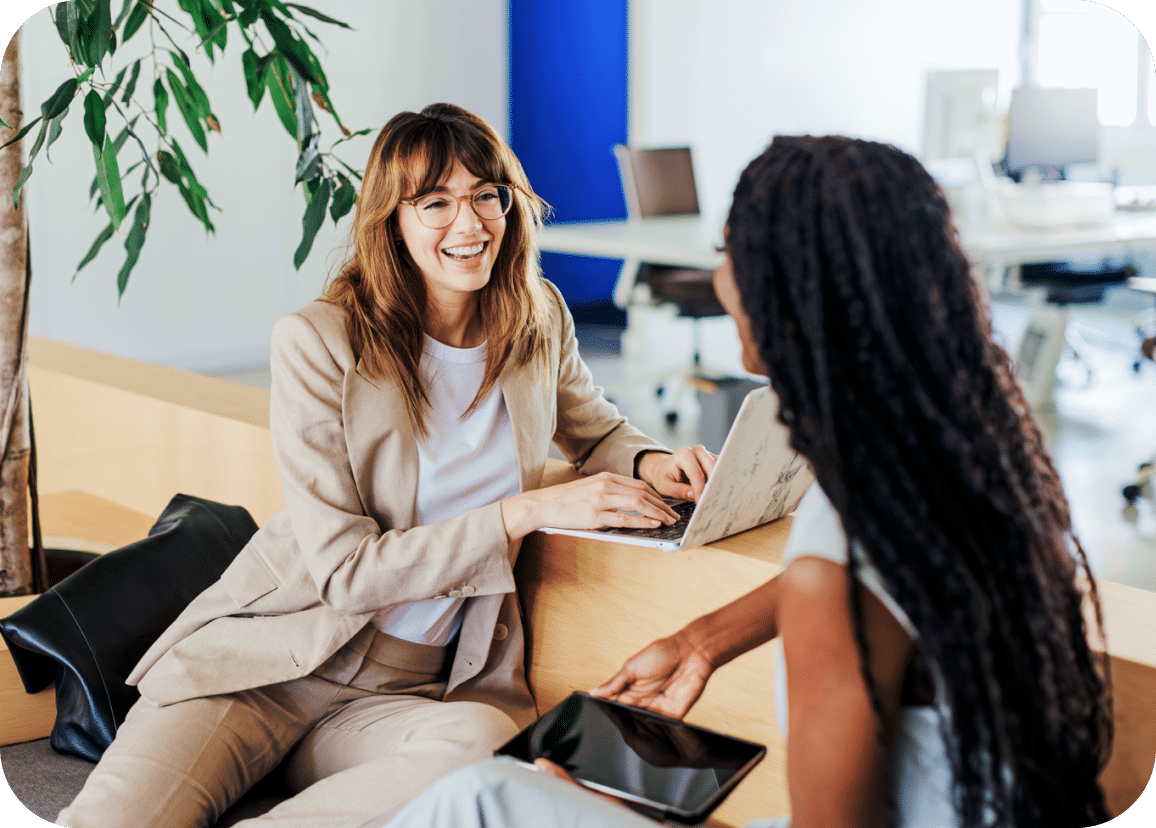 Two women sit and talk in a modern office, one smiling with a laptop and the other holding a tablet.