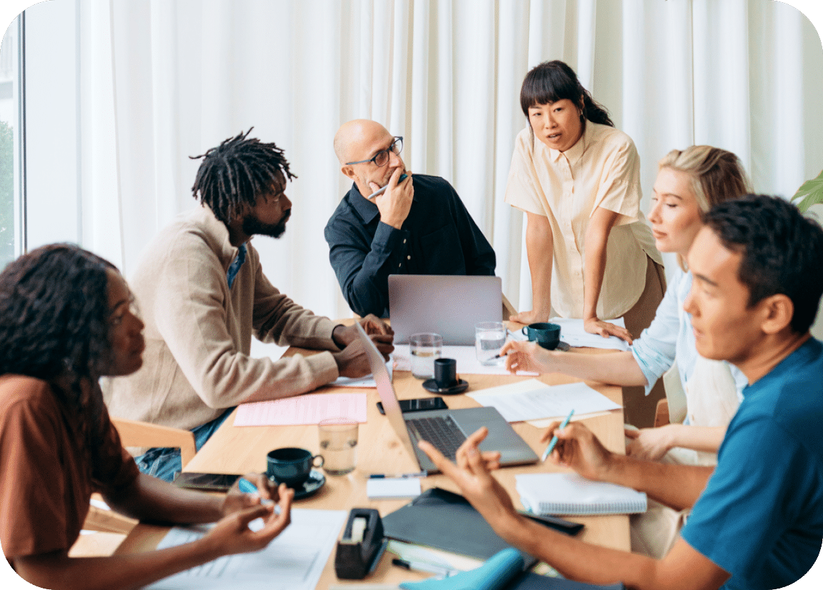 Six people sit around a table in a meeting, discussing and taking notes. Laptops, papers, and coffee cups are on the table. One woman stands, engaging with the group. The atmosphere appears collaborative and focused.