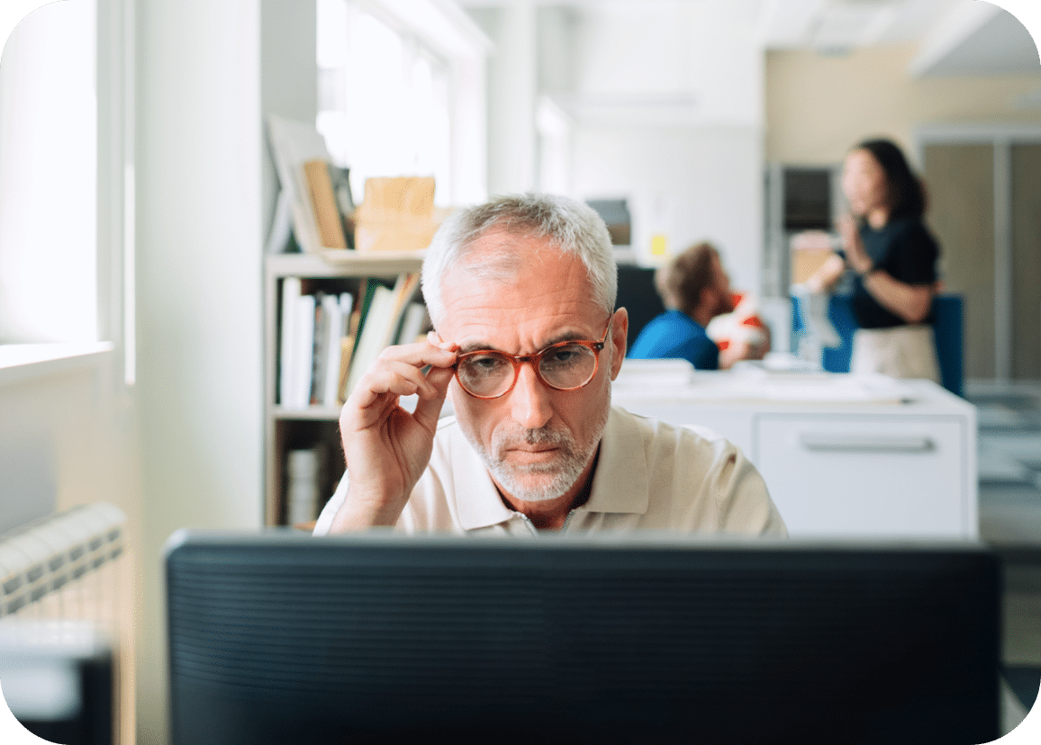 A middle-aged man with gray hair and glasses looks intently at a computer screen in an office, while two people talk in the background.