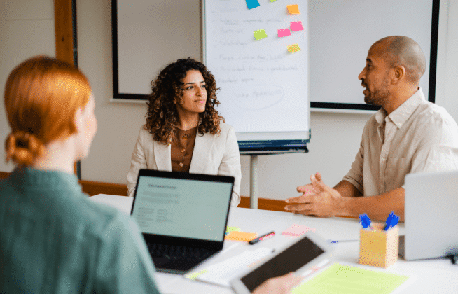 Three people having a discussion around a table with laptops, notebooks, and sticky notes; a whiteboard with notes and colorful sticky notes is visible in the background.