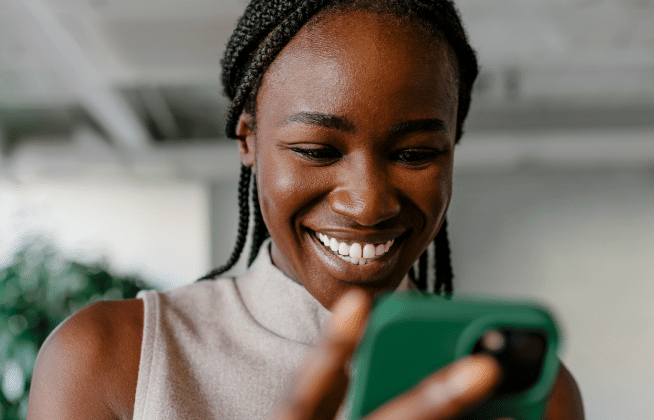 A woman with braided hair smiles while looking at a green phone, indoors with a blurred background.