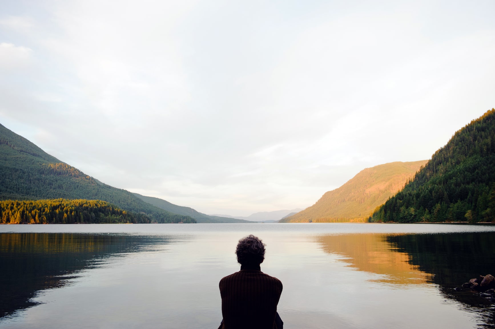A person sits by a calm lake surrounded by forested mountains under a partly cloudy sky, reflecting the landscape in the water at either sunrise or sunset.
