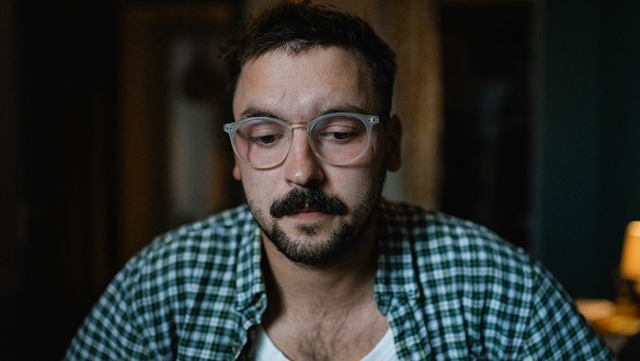 A man with glasses and a mustache, wearing a green plaid shirt, looks down with a thoughtful or concerned expression in a dimly lit room.