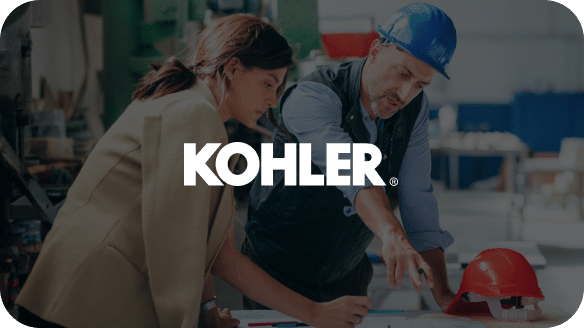A woman and a man in a blue hard hat review architectural plans at a workbench in an industrial setting. The man points at the plans while the woman takes notes. Features the Kohler logo on an overlay