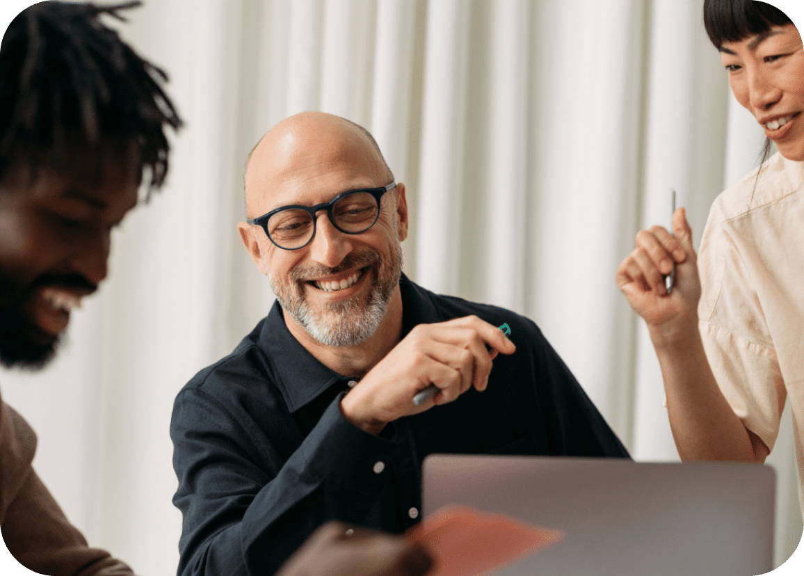 Three people are smiling and working together at a table. One person in glasses is holding a pen and looking at a laptop while the others engage in conversation, suggesting a friendly and collaborative work environment.