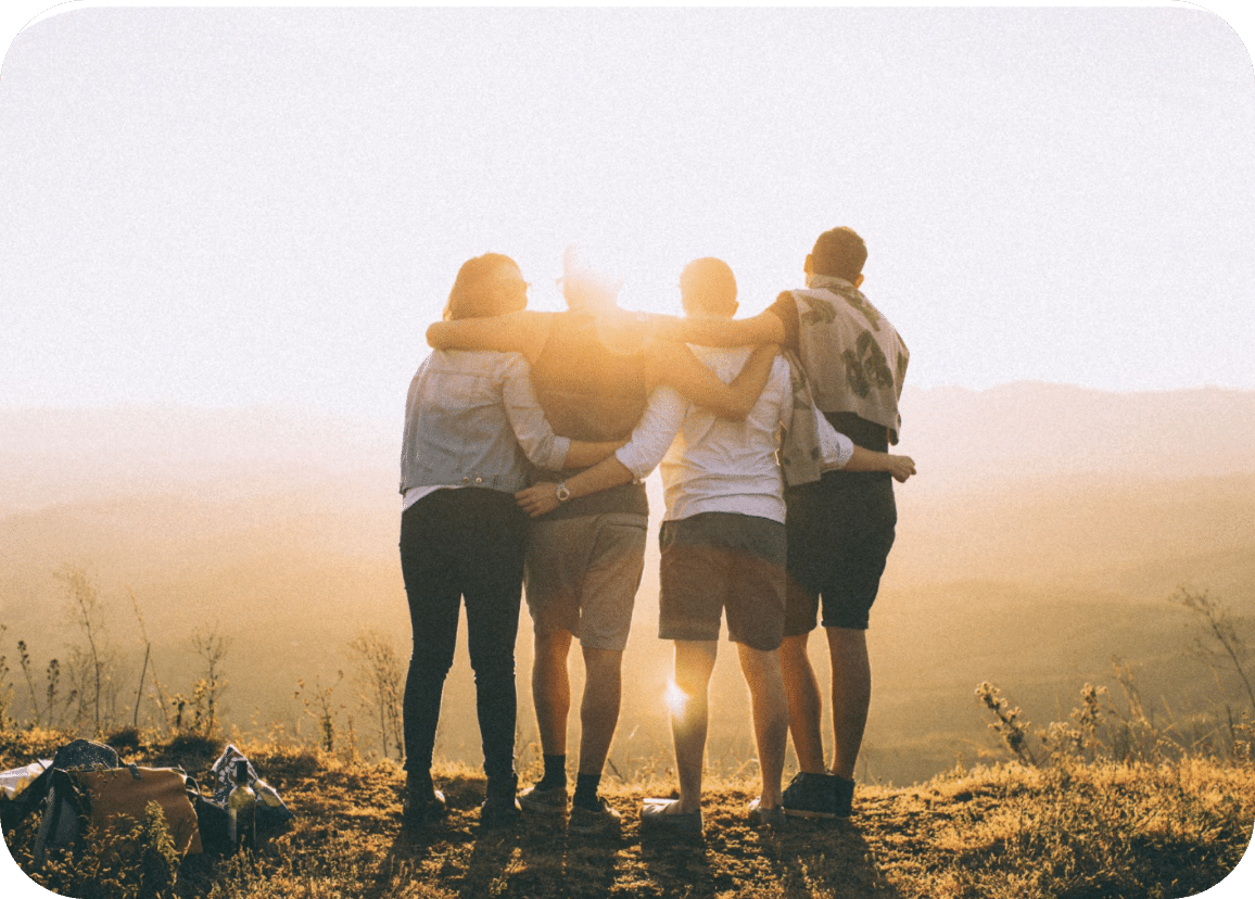 Four people stand side by side with arms around each other, facing a sunset over rolling hills, creating a warm, golden atmosphere.