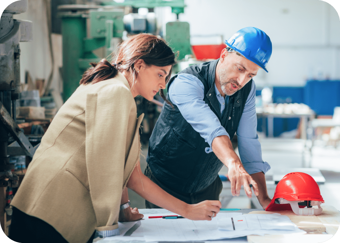 A woman and a man in a blue hard hat review architectural plans at a workbench in an industrial setting. The man points at the plans while the woman takes notes. A red hard hat sits nearby.