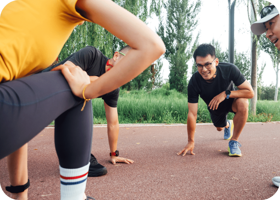 Three people stretch and warm up on an outdoor running track surrounded by greenery. One person lunges in the foreground while two others squat and smile, preparing for exercise.