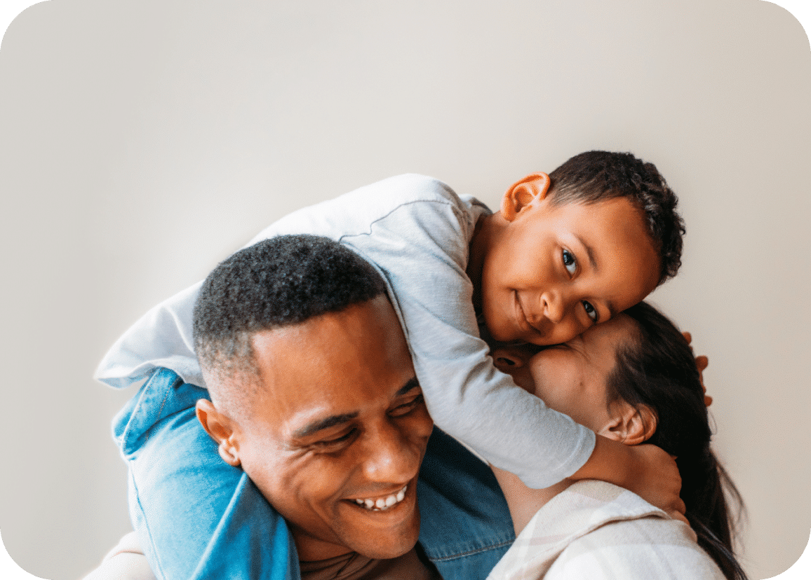 A smiling child lies on an adults shoulders, hugging another adult. All three appear happy, sharing a joyful moment together against a plain background.