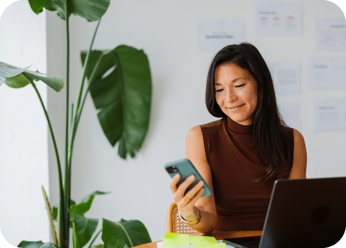 A woman sits at a desk with a laptop, looking at her phone and smiling. There are plants nearby and papers on the wall behind her.