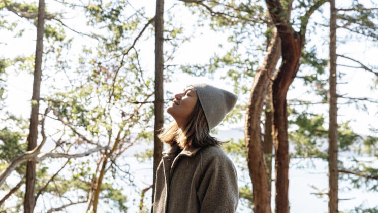 A person wearing a gray coat and beanie stands among tall trees, eyes closed and face tilted up toward the sunlight, surrounded by a forest with light streaming through the branches.