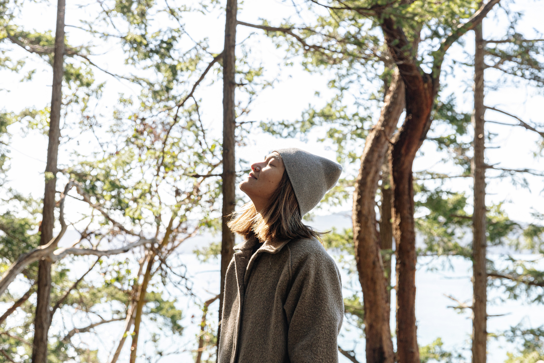 A person wearing a gray coat and beanie stands among tall trees, eyes closed and face tilted up toward the sunlight, surrounded by a forest with light streaming through the branches.
