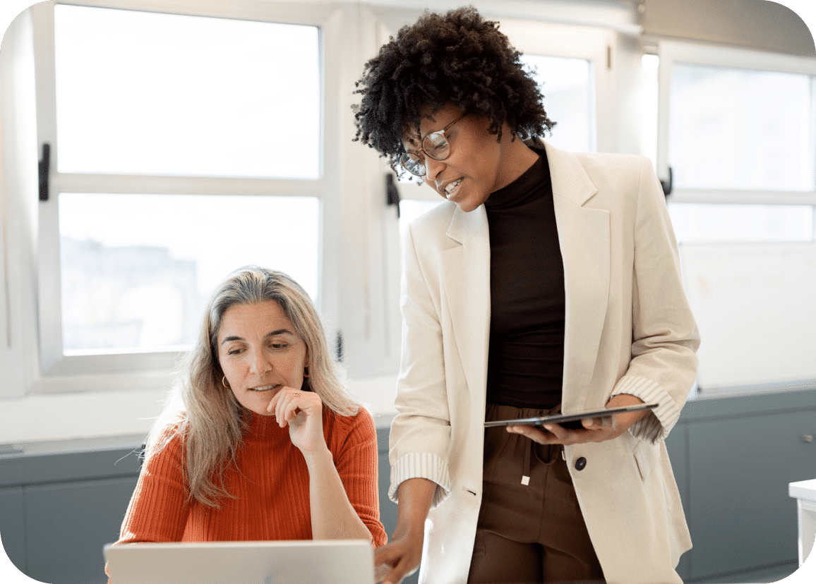 Two women in an office work together at a desk. One, seated in an orange sweater, looks at a laptop while the other, standing in a cream blazer, holds a tablet and gestures toward the screen.