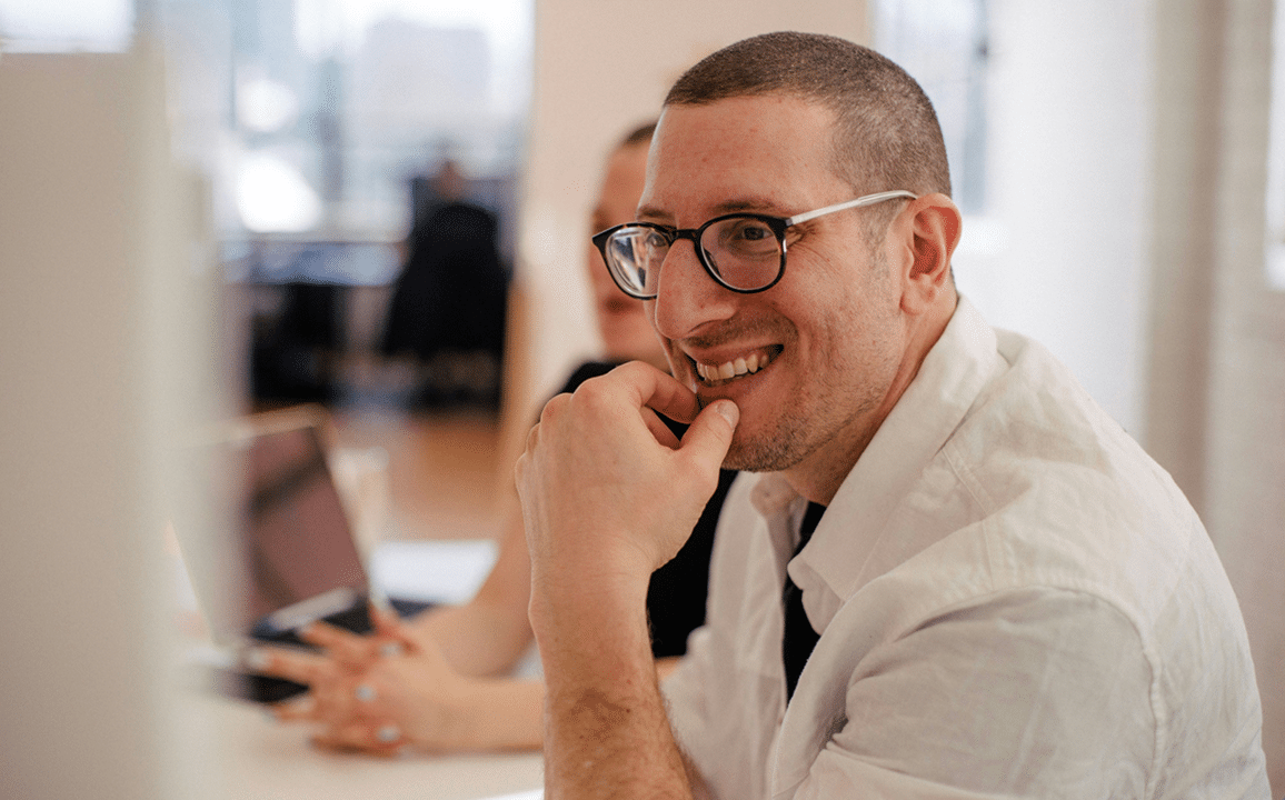 A man in glasses and a white shirt smiles while sitting at a desk with another person and a laptop in a bright office setting.