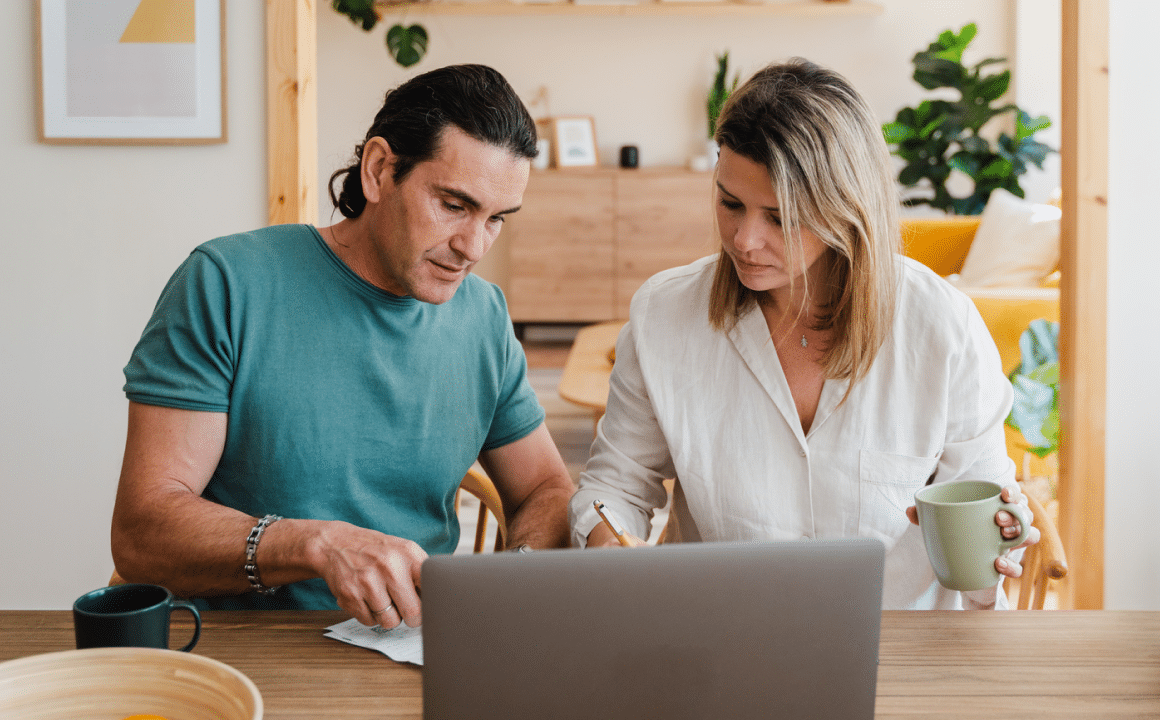A man and woman sit at a table, looking at papers and a laptop. The woman holds a mug, and both appear focused. They are in a bright home setting with plants and wooden furniture.