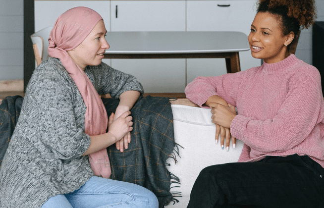 Two women sit on a couch having a conversation. One wears a pink headscarf and gray sweater, while the other wears a pink sweater and black pants. They appear to be engaged in a friendly discussion indoors.