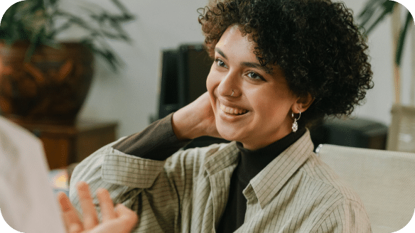 A person with short curly hair smiles and touches their neck while sitting indoors, engaged in conversation with someone out of frame.