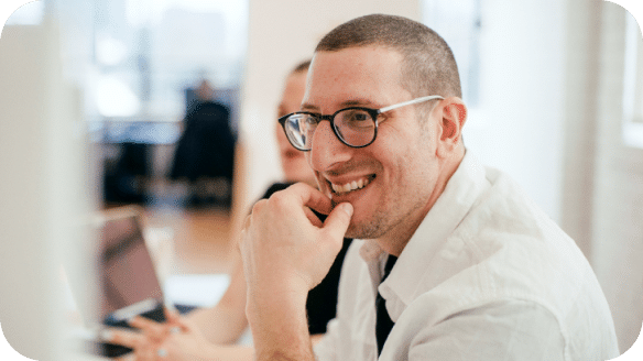 A man wearing glasses and a white shirt smiles while sitting at a desk, with a blurred person and laptop in the background.