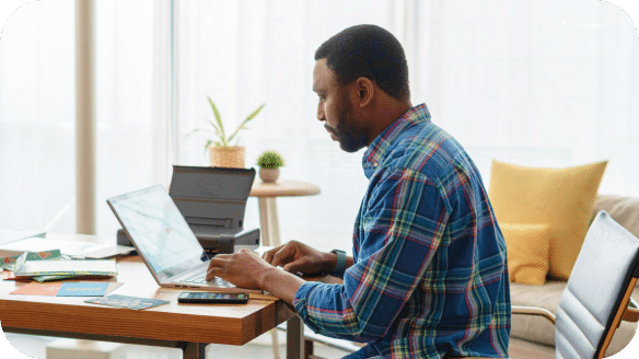 A man in a plaid shirt types on a laptop at a wooden desk in a bright, modern living room, with papers, a phone, and a potted plant nearby.