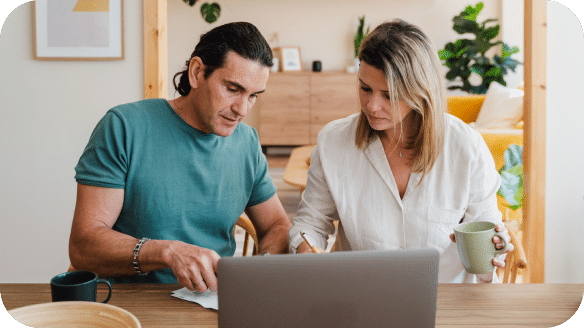 A man and woman sit at a table with a laptop, reviewing papers together. The woman holds a mug, and both look focused. The background shows a cozy, well-lit living room with plants and stylish decor.