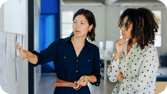 Two women stand by a whiteboard covered in papers; one woman points at the board while explaining something, and the other listens thoughtfully with her hand on her chin.