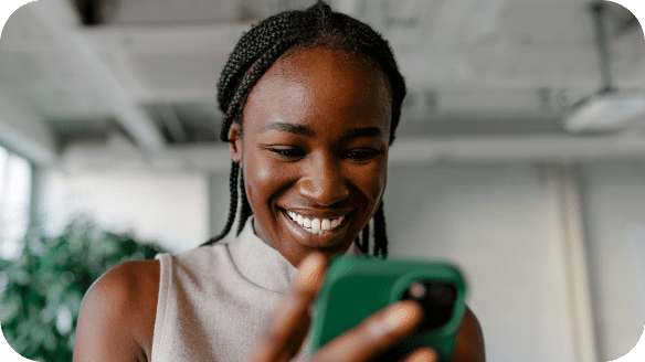 A woman with braided hair smiles while looking at her phone, indoors with a blurred background.