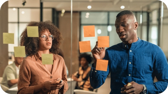 Two people stand in front of a glass wall with sticky notes, discussing ideas in a modern office setting. Other colleagues are blurred in the background.