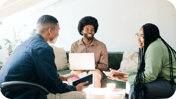 Three people sit together on a couch, smiling and talking. One holds a laptop, another holds a tablet, and the third has a notebook. They appear to be having a relaxed group discussion in a bright room.